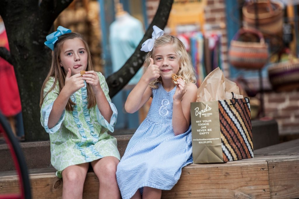 Two young girls sitting on a wood bench eating cookies in the Savannah City Market courtyard.